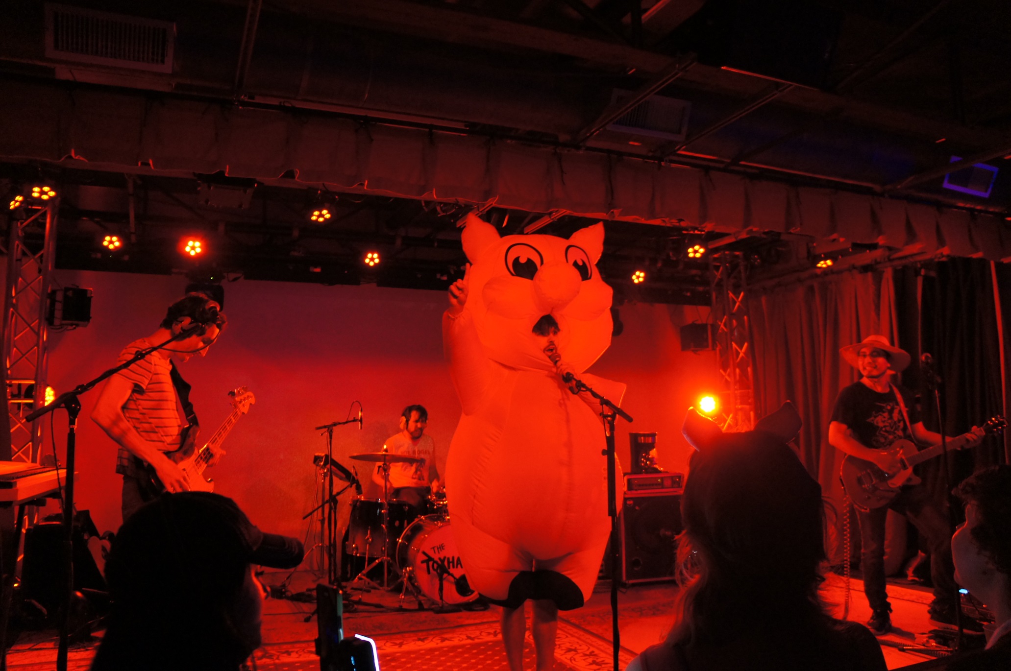 Heavily red-tinted image of the Toxhards on stage, with Alan Macchiarolo playing the drums, Emerson Harrison playing the bass, Matt Garcia playing the guitar, and Casey Donovan, wearing a giant inflatable hog suit, singing lead and pointing at the sky.